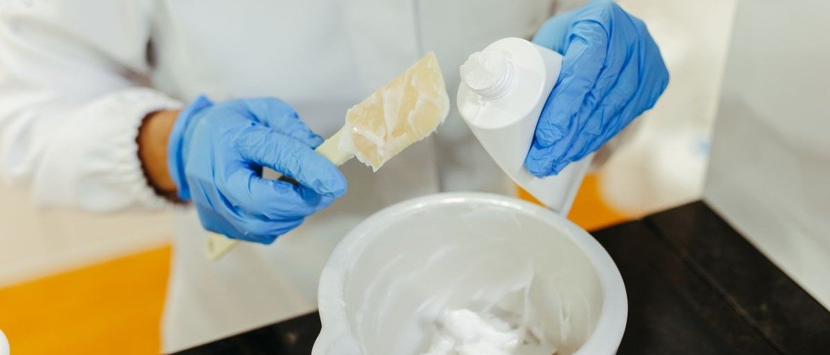 A formulator in blue gloves adding a waxy ingredient to a white cream base in a mixing bowl